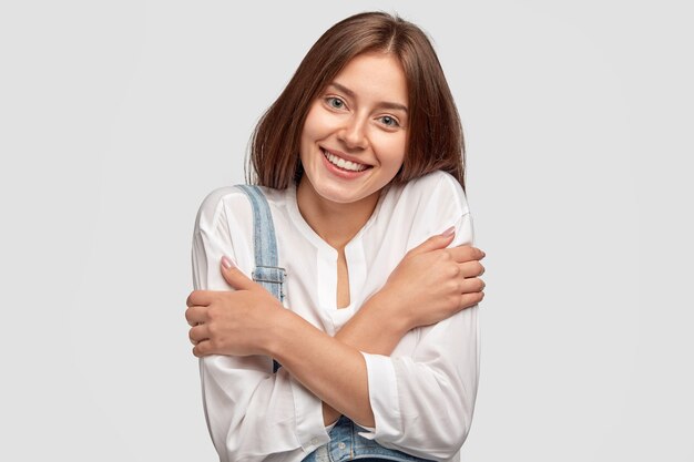 Une fille gaie timide s'embrasse, exprime des émotions positives, se sent heureuse de recevoir des compliments ou des éloges, vêtue d'une chemise élégante