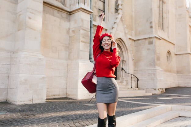 Fille galbée en bottes hautes en cuir posant avec les mains près du vieux bâtiment