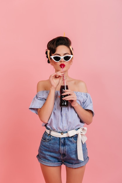 Photo gratuite fille galbée en short en jean et chemisier rayé, boire des boissons. photo de studio de pin-up à lunettes de soleil.