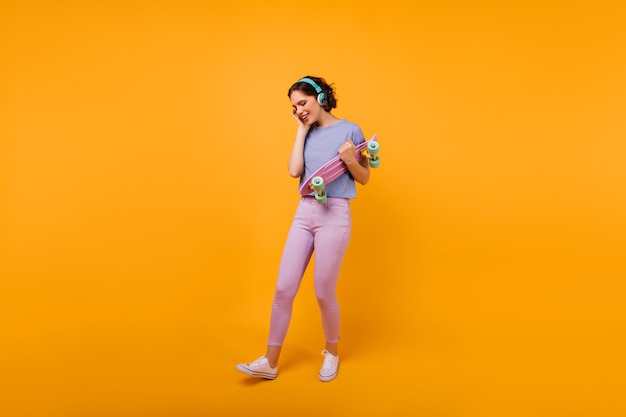 Fille galbée en tenue décontractée colorée, écouter de la musique dans les écouteurs. Modèle féminin blithesome avec coupe de cheveux courte tenant une planche à roulettes.