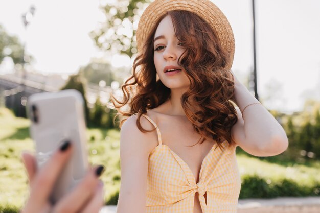 Fille de gingembre inspirée prenant une photo d'elle-même avec une expression de visage intéressée. Photo extérieure d'une magnifique femme aux cheveux roux utilisant un smartphone blanc pour selfie.