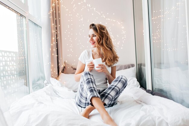 Fille glamour avec un sourire heureux se détendre dans le bonjour. Modèle féminin positif buvant du thé chaud.