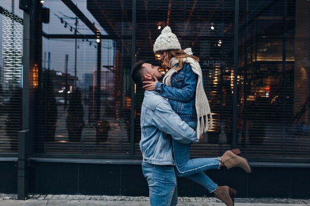 Fille gracieuse en écharpe tricotée et bottes à talons hauts s'amusant pendant la date. Portrait en plein air de mec européen tenant sa petite amie sur la rue urbaine.