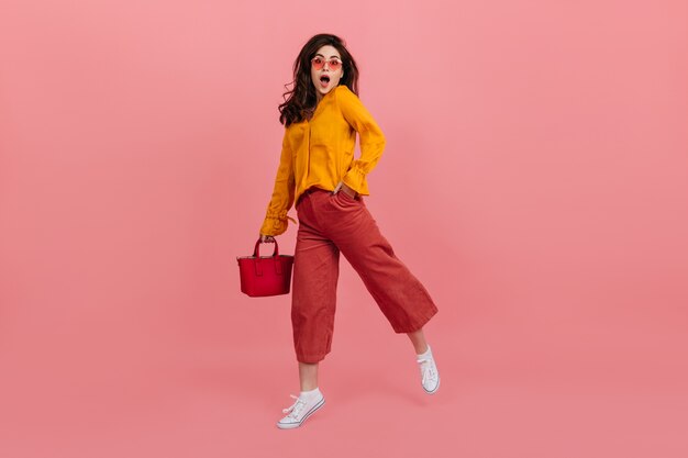 Fille guillerette dans des lunettes élégantes regarde avec étonnement, marchant sur le mur rose. Brunette en jupe-culotte et chemisier orange posant avec sac à main rouge.