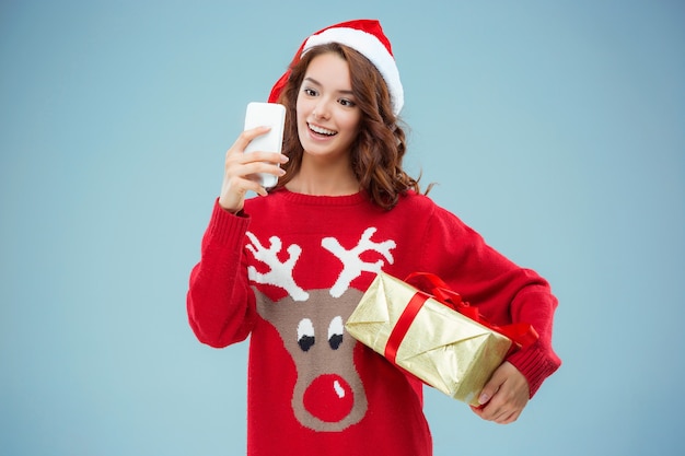 Fille habillée en bonnet de Noel avec un cadeau de Noël et un téléphone. Elle prend une photo de selfie. Concept de vacances avec fond bleu.