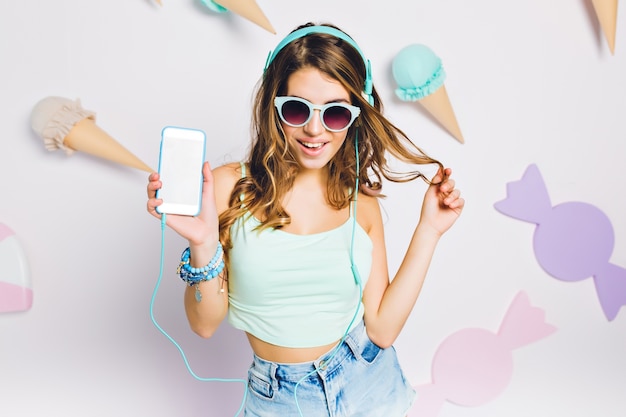 Fille heureuse en accessoires bleus écoutant de la musique dans sa chambre, tenant un smartphone et jouant avec les cheveux. Portrait d'élégante jeune femme dans des verres avec une coiffure incroyable bouclés posant sur un mur violet.