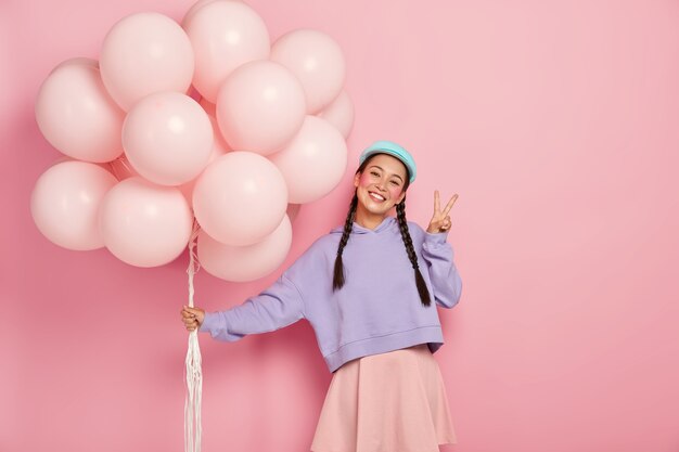 Une fille heureuse accueille des amis lors d'une fête de ballon, a deux tresses, porte un pull et une jupe violets, fait un geste de paix, se tient contre le mur rose