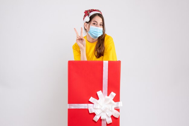 Fille heureuse avec Bonnet de Noel faisant signe de la victoire debout derrière un grand cadeau de Noël sur blanc
