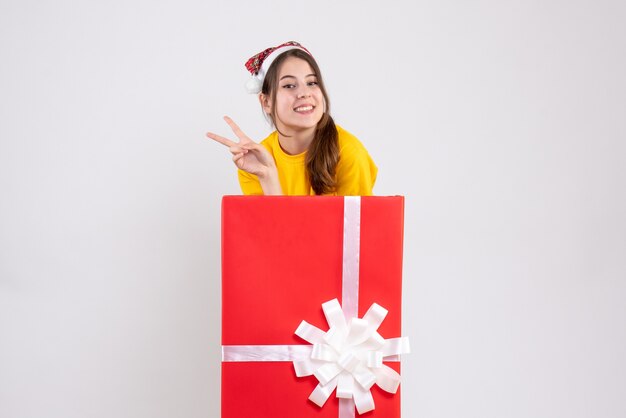 Fille heureuse avec Bonnet de Noel faisant signe de la victoire debout derrière un grand cadeau de Noël sur blanc