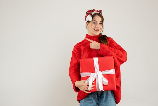 fille heureuse avec bonnet de noel tenant présent doigt pointant quelque chose debout sur blanc