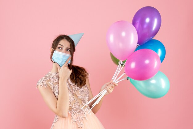 fille heureuse avec chapeau de fête et masque médical tenant des ballons colorés mettant la main sur son menton rose