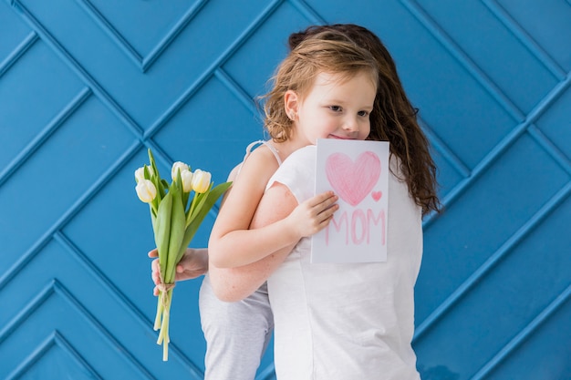 Photo gratuite fille heureuse étreignant sa mère avec des fleurs et une carte de voeux sur fond bleu