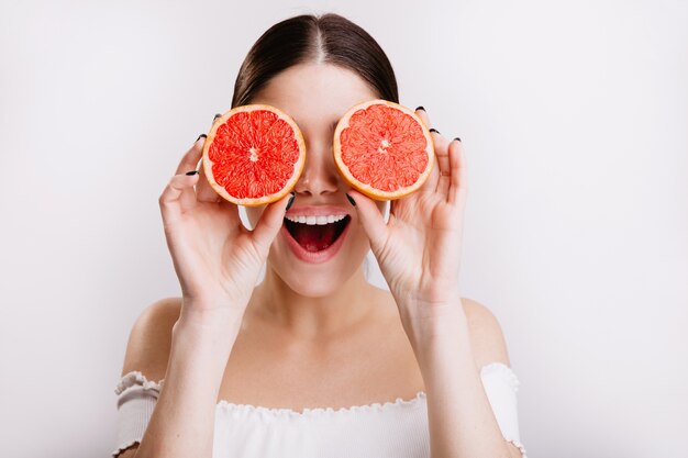 Fille heureuse avec une expression faciale émotionnelle positive couvre ses yeux d'oranges, posant sur un mur isolé.