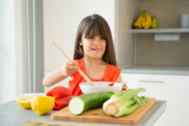 Fille heureuse en jetant la salade dans un bol avec une grande cuillère en bois. Adorable enfant apprenant à cuisiner des légumes pour le dîner, posant, sortant la langue. Apprendre à cuisiner le concept
