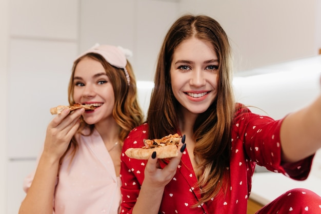 Fille heureuse avec manucure noire faisant selfie pendant que son amie mangeait de la pizza. Portrait intérieur de deux sœurs se détendre avec de la restauration rapide.