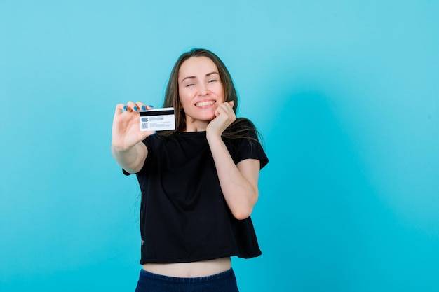 Une fille heureuse montre une carte de crédit à la caméra et tient l'autre main sur la joue sur fond bleu
