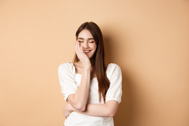 Fille heureuse et romantique riant avec les yeux fermés touchant le visage et ressentant de la joie debout sur un ba beige ...