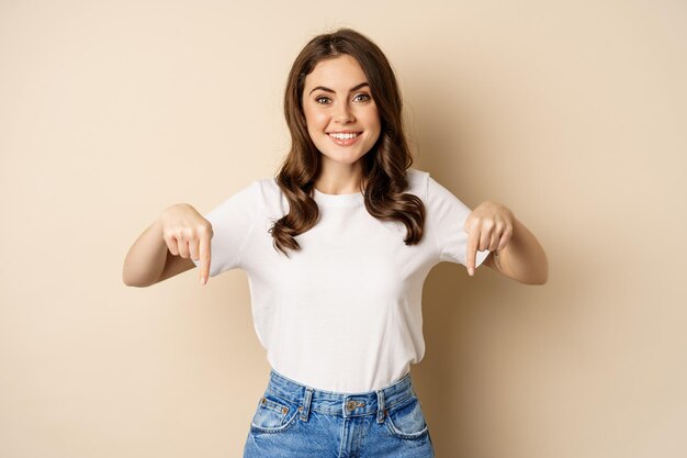 Fille heureuse souriante pointant les doigts vers le bas et montrant la bannière, le logo de l'entreprise ci-dessous, cliquez sur le lien, debout sur fond beige