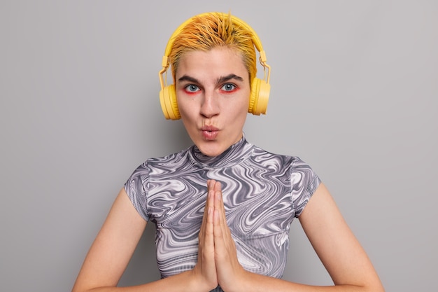 Photo gratuite une fille hipster aux cheveux jaunes surprise appuie sur les paumes ensemble croit en quelque chose de bien garde les lèvres arrondies a l'air étonnamment a un maquillage brillant écoute une piste audio isolée sur un mur gris