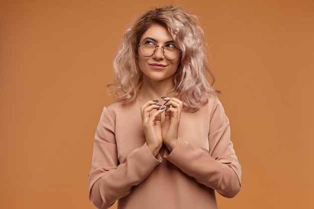 Fille de hipster sournois planifiant une farce ou un truc maléfique, serrant les mains et souriant mystérieusement. Pensive jeune femme rusée en lunettes ayant un plan délicat à l'esprit