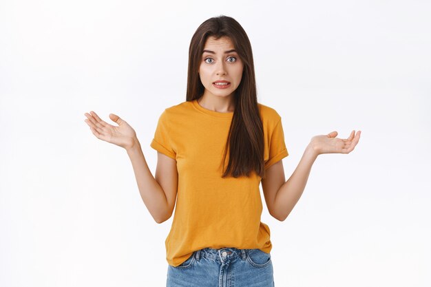 Une fille idiote perplexe, maladroite et embarrassée fait une erreur en disant oups, en haussant les épaules et en tenant les mains écartées, ne peut pas expliquer comment la situation s'est produite, souriante inquiète, debout sur fond blanc