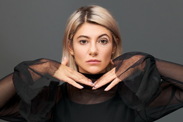 Fille incroyable avec une coiffure teinte blonde, un anneau dans le nez et un maquillage élégant allant faire la fête avec des amis à la fête d'anniversaire portant un chemisier noir avec des fusées éclairantes, se tenant la main sous le menton