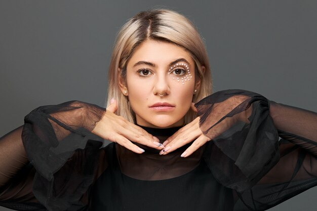 Fille incroyable avec une coiffure teinte blonde, un anneau dans le nez et un maquillage élégant allant faire la fête avec des amis à la fête d'anniversaire portant un chemisier noir avec des fusées éclairantes, se tenant la main sous le menton