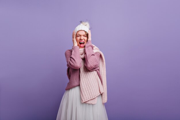 Fille incroyable en longue écharpe tricotée hurlant les yeux fermés. Magnifique dame européenne dans des vêtements d'hiver élégants posant sur un mur violet