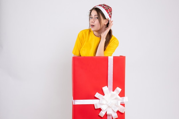 Photo gratuite fille indiscrète avec bonnet de noel debout derrière un gros cadeau de noël sur blanc