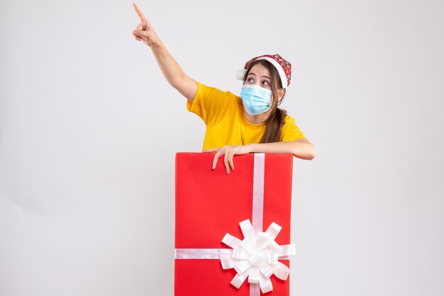 Fille indiscrète avec Bonnet de Noel montrant quelque chose debout derrière un gros cadeau de Noël sur blanc