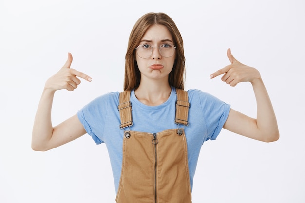 Fille insécurisée triste et sombre dans des lunettes pointant sur elle-même ou un logo, ressentant des regrets ou de la colère