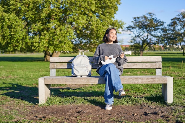 Photo gratuite une fille insouciante est assise sur un banc dans un parc avec un ukulélé joue et chante à l'extérieur par une journée ensoleillée