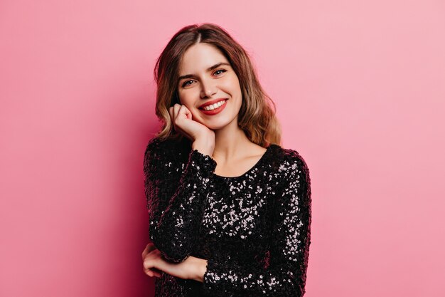 Fille insouciante en robe noire souriant. Modèle féminin élégant posant sur un mur rose.