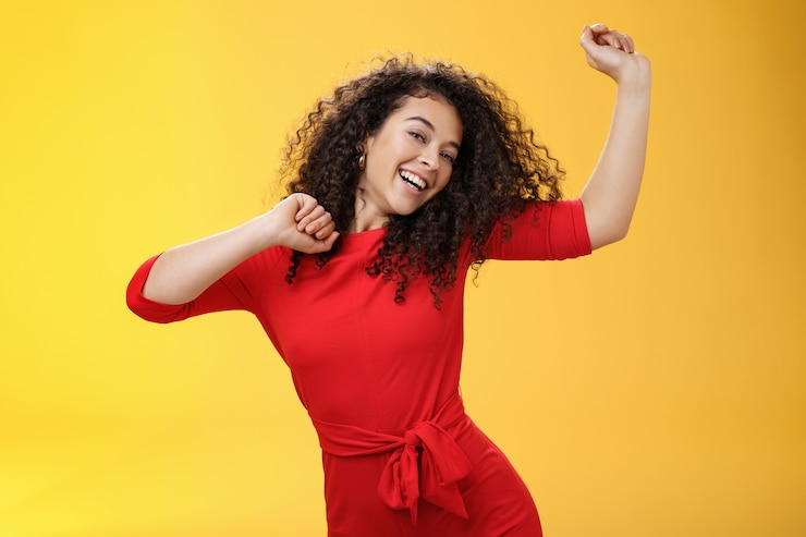 Photo gratuit fille insouciante se sentant exaltée et joyeuse dansant en robe rouge levant les mains en inclinant joyeusement la tête et souriant largement à la caméra comme profitant de vacances, célébrant les vacances sur fond jaune.
