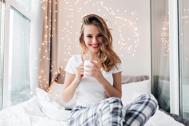 Fille insouciante avec un sourire joyeux posant dans son lit dans sa chambre. Portrait intérieur de femme blonde débonnaire tenant une tasse de thé.