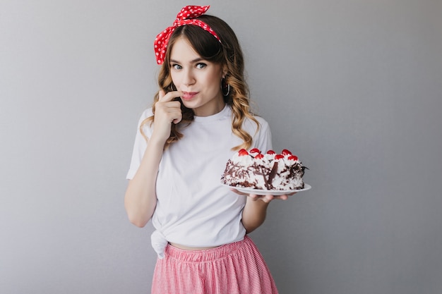 Fille inspirée aux cheveux ondulés dégustant un gâteau d'anniversaire. modèle féminin enchanteur avec tarte posant.