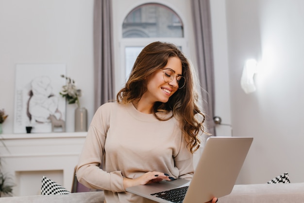 Fille intelligente insouciante en chemise beige posant avec un sourire romantique, travaillant avec ordinateur