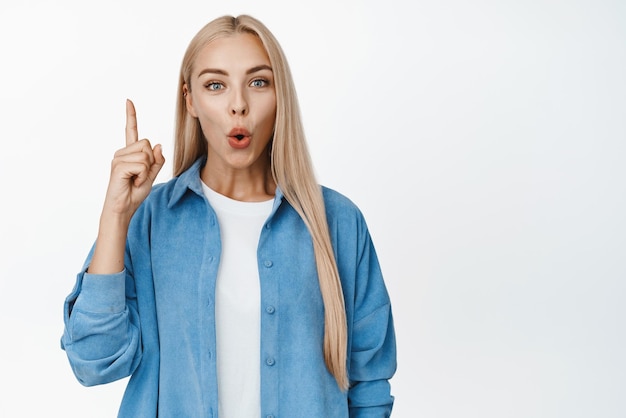 Fille intriguée pointant le doigt vers le haut dire wow l'air impressionné debout sur fond blanc