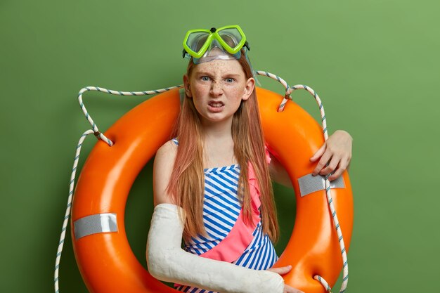 Une fille irritée ne peut pas nager à cause d'une fracture du bras, porte un plâtre, pose avec des lunettes de natation et un anneau gonflé, apprécie le repos d'été, recrée près de la mer, pose contre un mur végétal. Enfants, repos