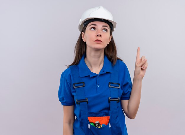 Fille jeune constructeur confus avec casque de sécurité blanc et uniforme bleu pointe vers le haut et lève les yeux sur fond blanc isolé avec espace de copie