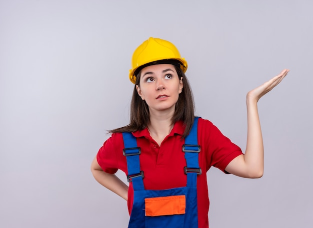 Fille jeune constructeur confus tient la main vide et lève les yeux sur fond blanc isolé avec espace copie