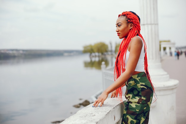 Une fille jeune et élégante à la peau foncée avec des dreads rouges marchant dans le parc de l&#39;été près de la rivière