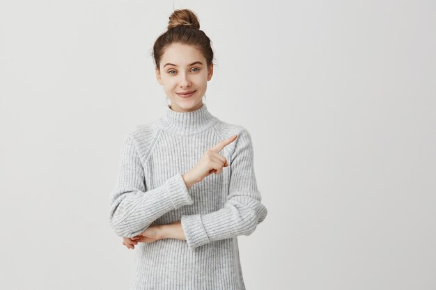 Fille joyeuse aux cheveux bruns étant heureuse et pointant le doigt sur le côté. Artiste féminine présentant son travail avec un large sourire. Concept de plaisir