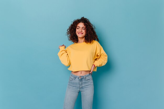 Fille joyeuse aux lèvres rouges danse sur l'espace bleu. Instantané de femme en sweat-shirt court et jeans.