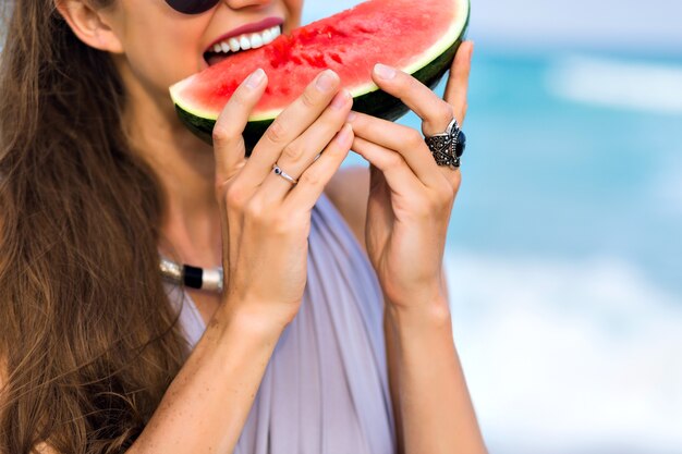 Fille joyeuse aux longs cheveux châtain clair mordant une pastèque. Close-up portrait de modèle féminin excité dans de grandes lunettes de soleil sombres appréciant la nourriture préférée avec le sourire.