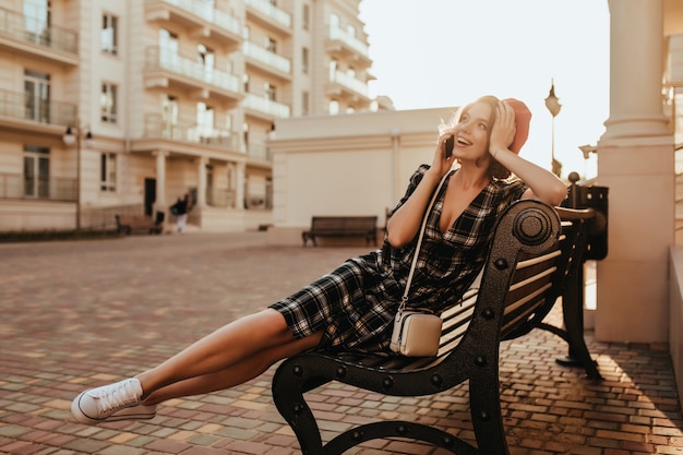 Fille joyeuse en baskets blanches, assis sur un banc en soirée. Photo extérieure d'une incroyable dame brune parlant au téléphone dans la rue.