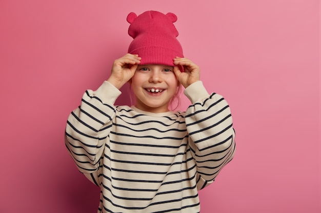 Une fille joyeuse insouciante met un chapeau rose avec des oreilles, sourit amicalement, se satisfait de la nouvelle tenue, parle avec son meilleur ami, porte un pull rayé surdimensionné, exprime de bonnes émotions, pose seule à l'intérieur