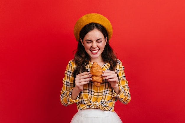 Fille joyeuse regarde un croissant fraîchement sorti du four en prévision. Portrait de dame en béret orange et jupe blanche sur mur rouge.