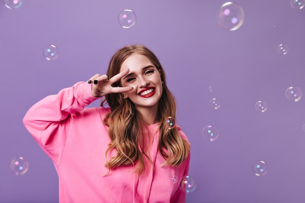 Fille joyeuse en sweat à capuche rose montrant un signe de paix sur un mur violet avec des bulles