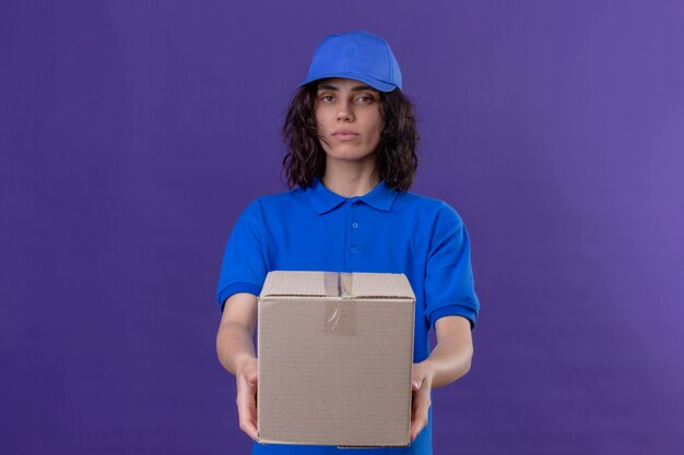Fille de livraison en uniforme bleu et cap étirant le paquet fort avec une expression confiante sérieuse debout sur violet isolé